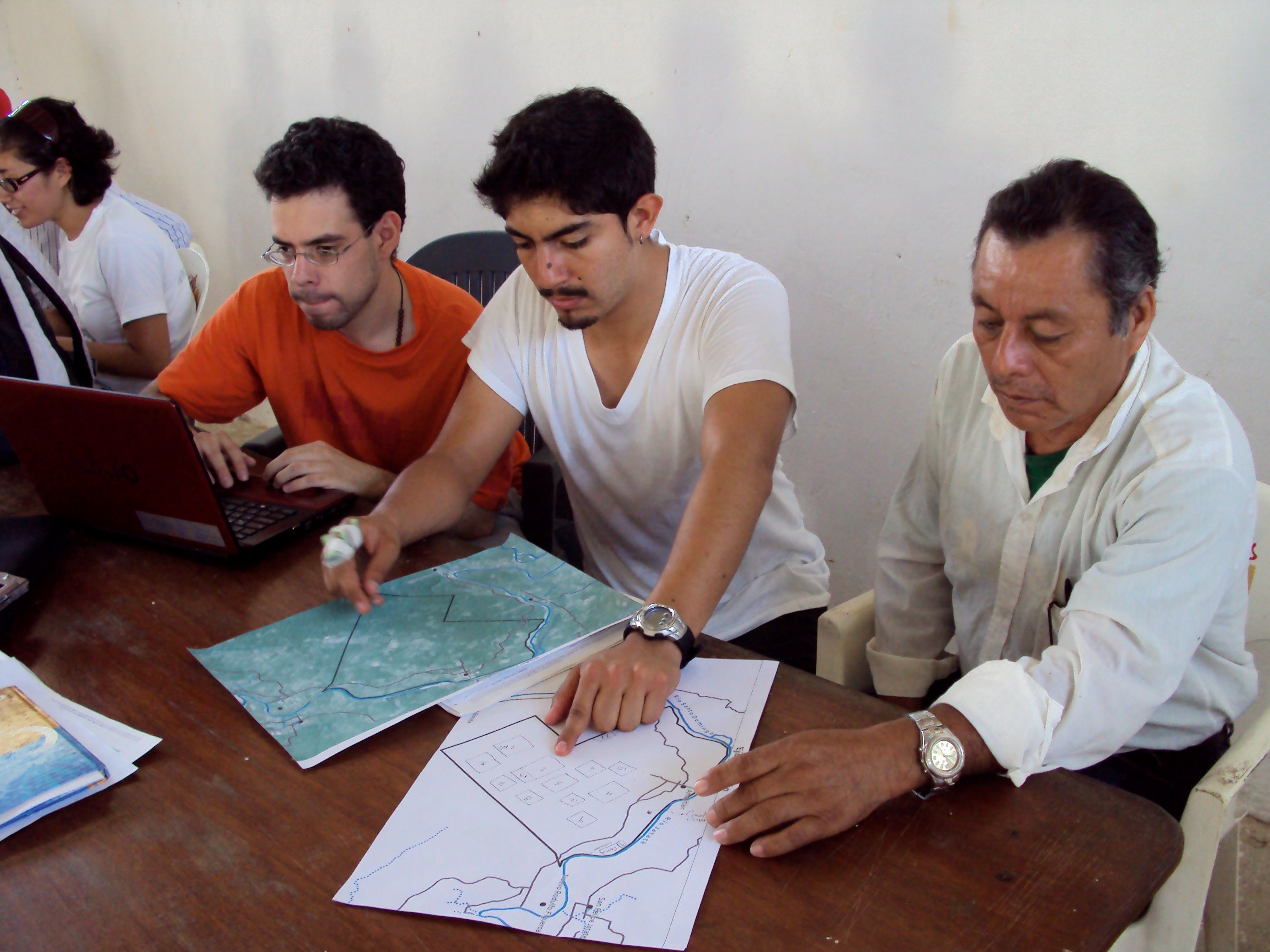 Three men, Talib among them, are seated at a table and looking at maps in front of them.
