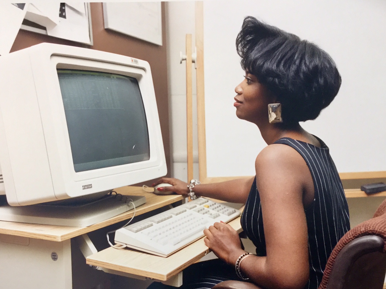 Cozette Parker, with elegant hair, earrings, and dress, is sitting at a desk, facing an old-style, massive-sized computer from the 1990s, her hand on the mouse, and a cheerful smile on her face.