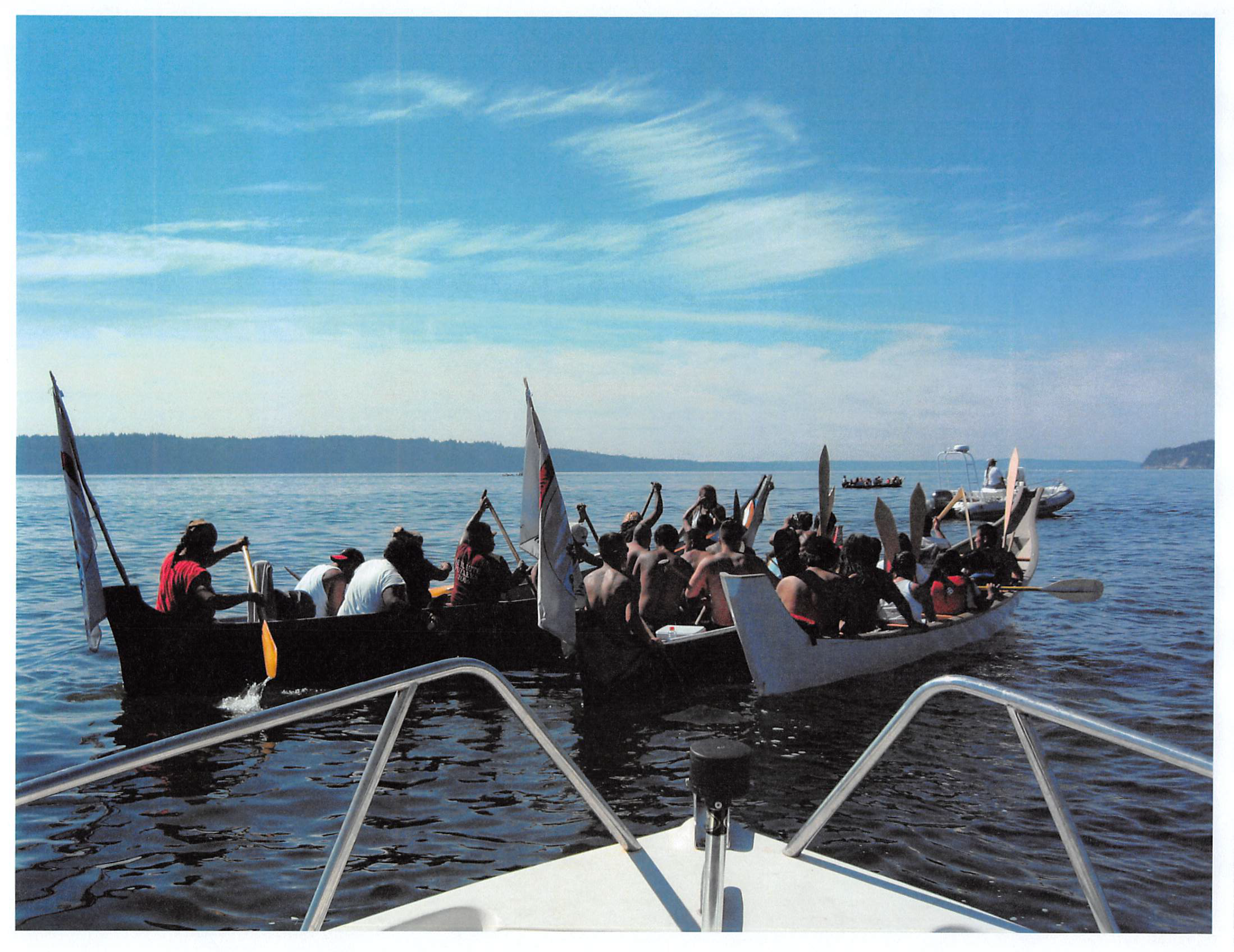 About 30 members of the Quileute tribe are seated in 2-3 canoes, setting off on a journey on blue waters and beneath a blue sky.