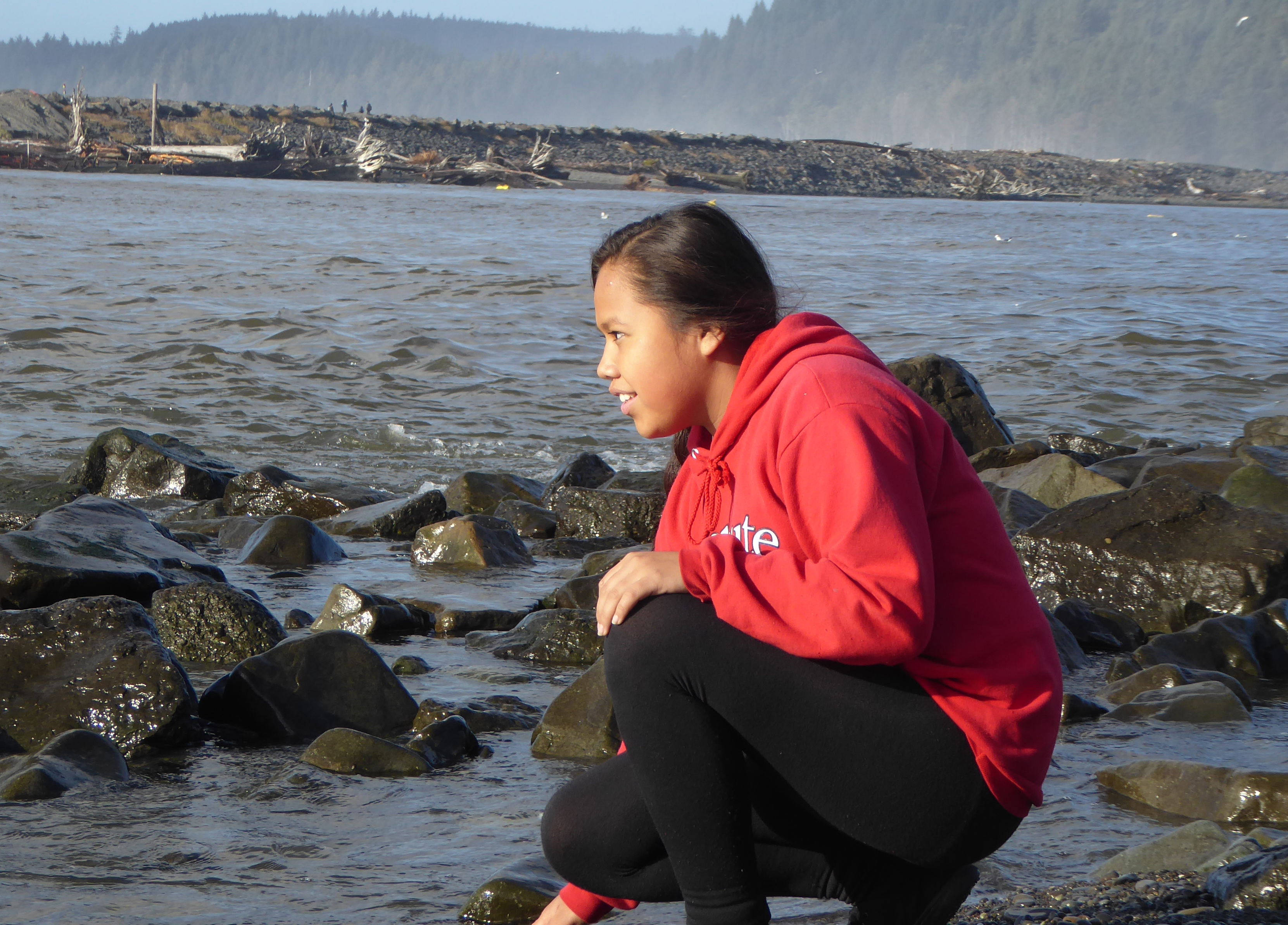 A student in a red sweater is perched on a rock in the middle of the water, with the water stretching in the background till it reaches forested mountains