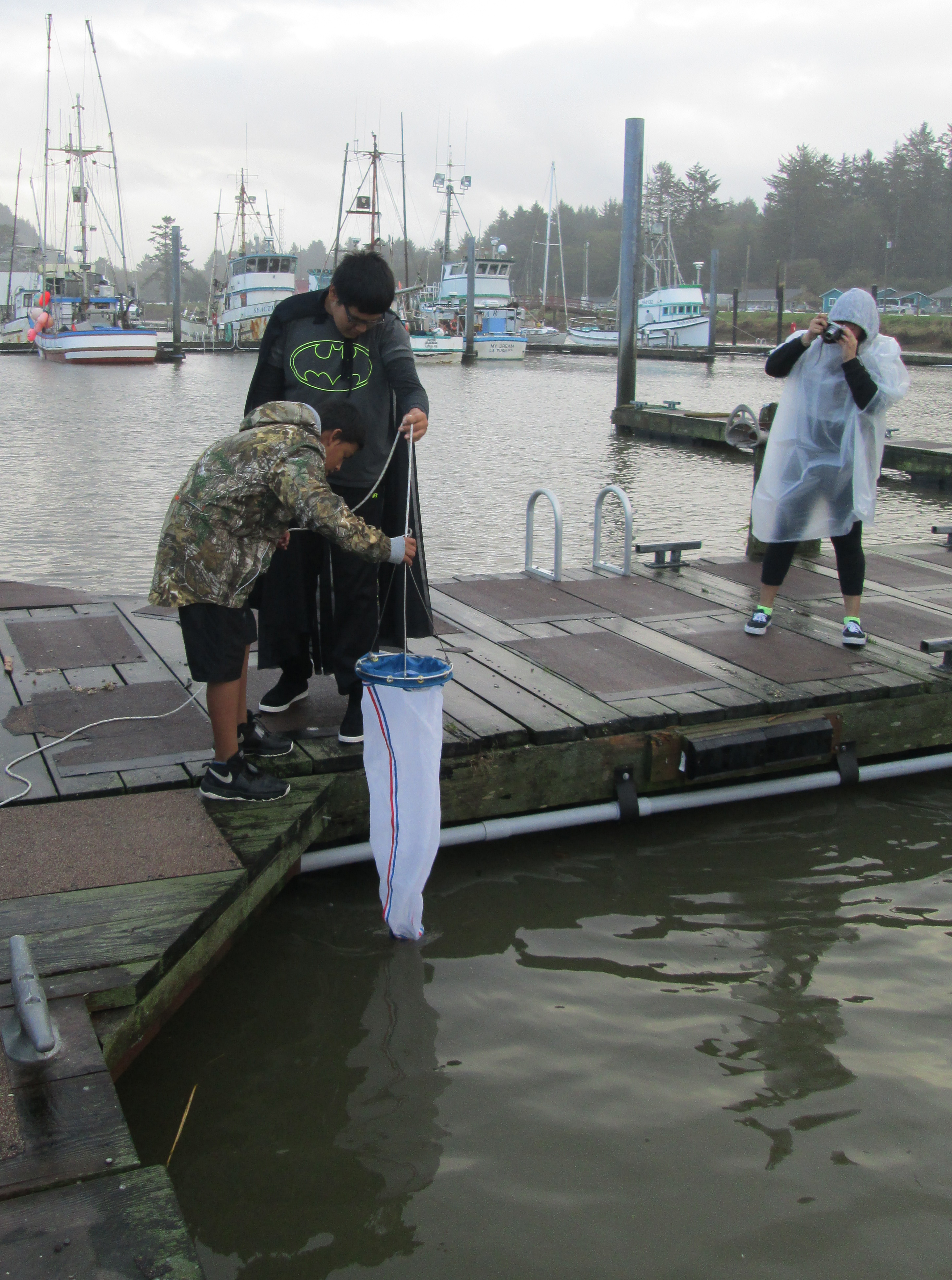 Two students stand on a damp pier on a cloudy day, small boats bobbing in the harbor in the background, and are pulling a plankton net out of the water, while a third person takes a photo of them. 