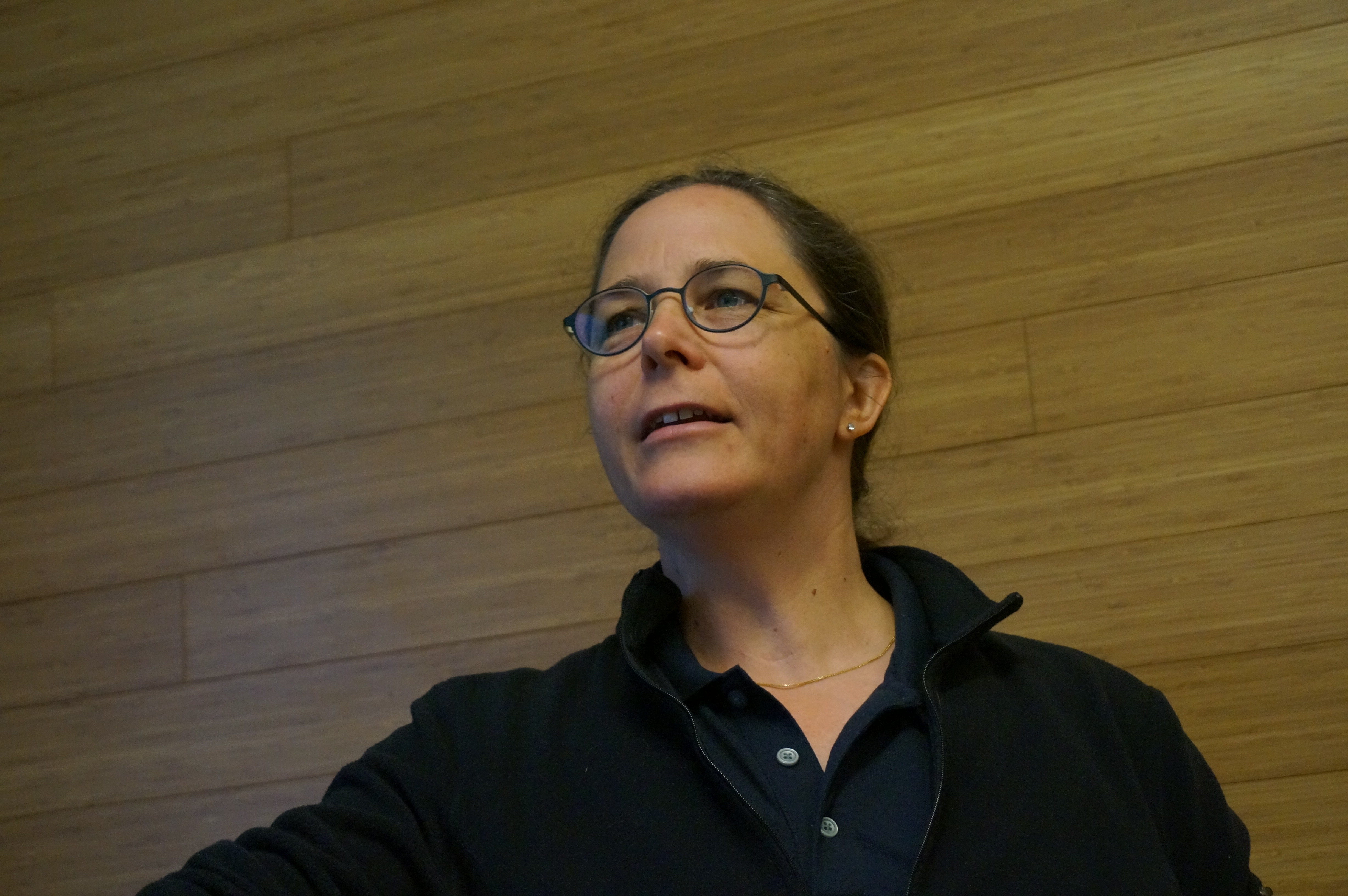 Annmarie Eldering is dressed in a black button-up shirt with a wood-paneled wall behind her, looking off to the side, a good-natured expression on her face. 