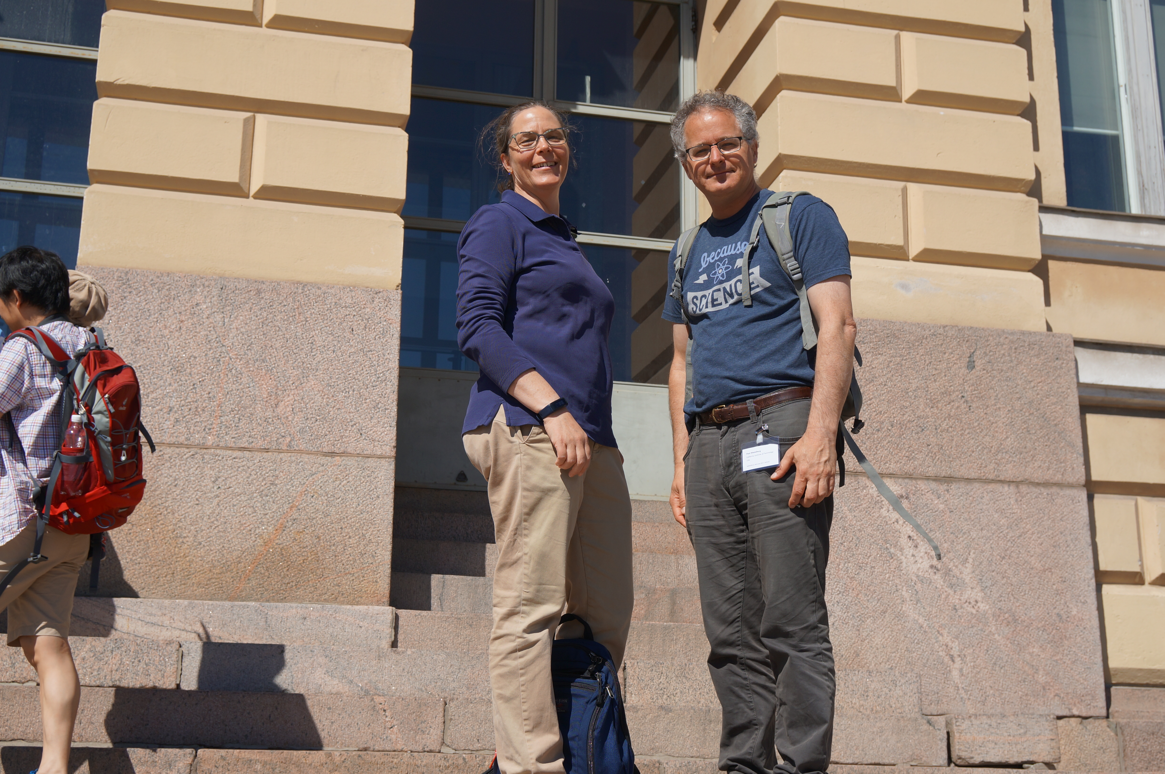 Annmarie and Paul Wennberg stand outdoors on the steps leading up to a large building, in bright sunshine, smiling down at the camera. 