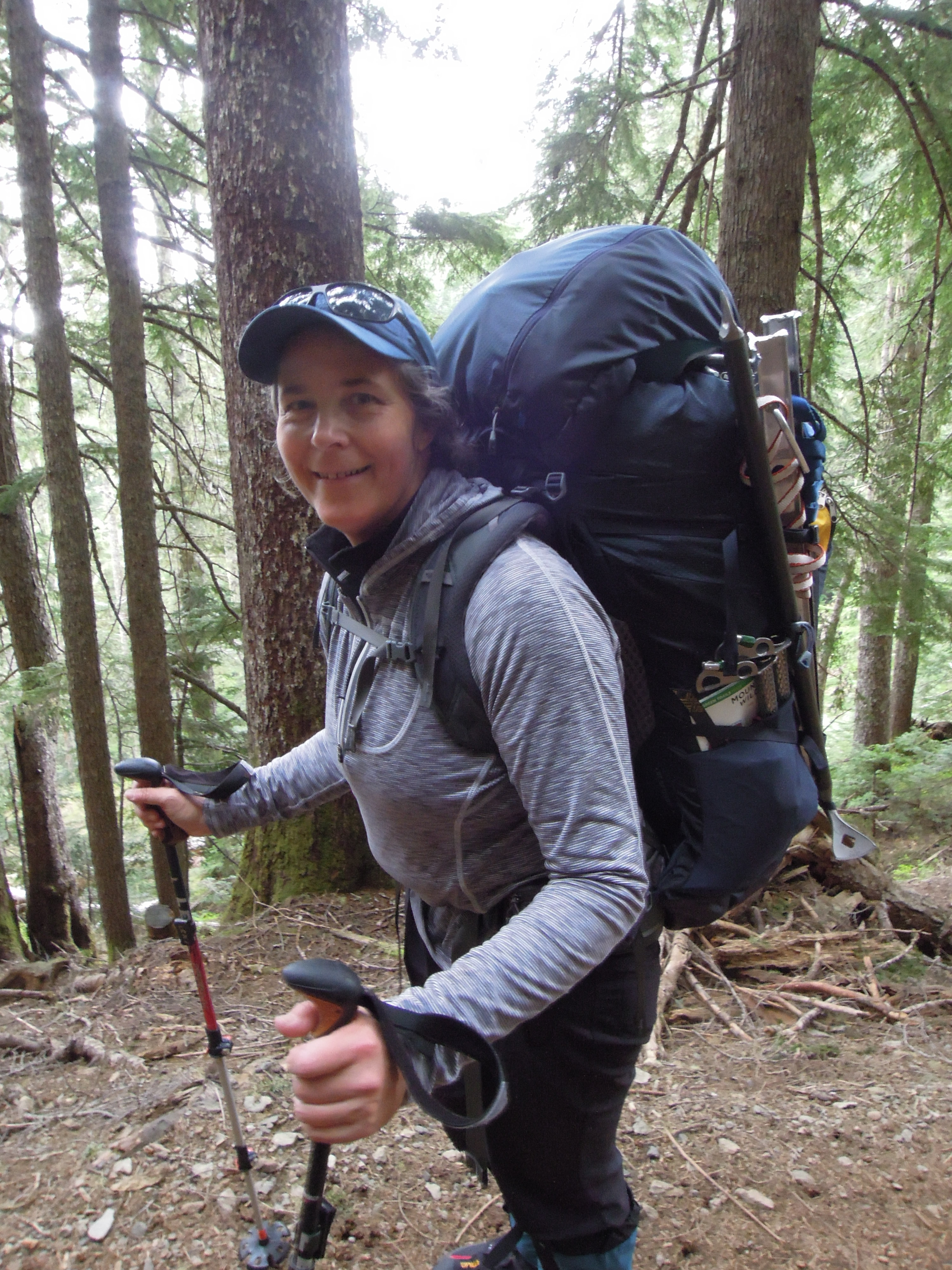 Annmarie hiking with a large backpack through a forest of tall conifers, wearing a baseball cap and smiling at the camera.