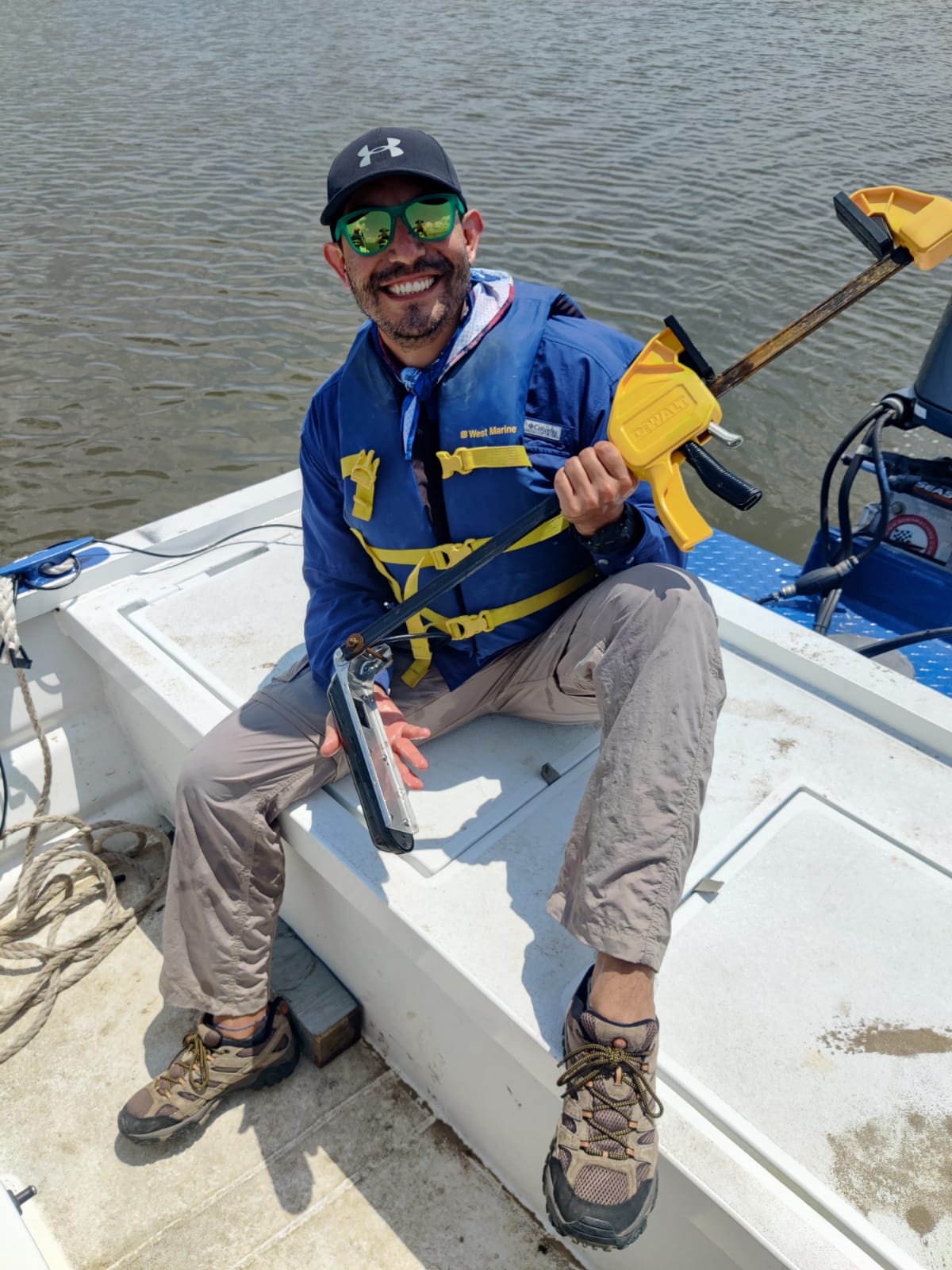 Talib is sitting on a boat, holding a long sonar instrument in his hand, wearing sunglasses and a hat, and smiling widely at the camera
