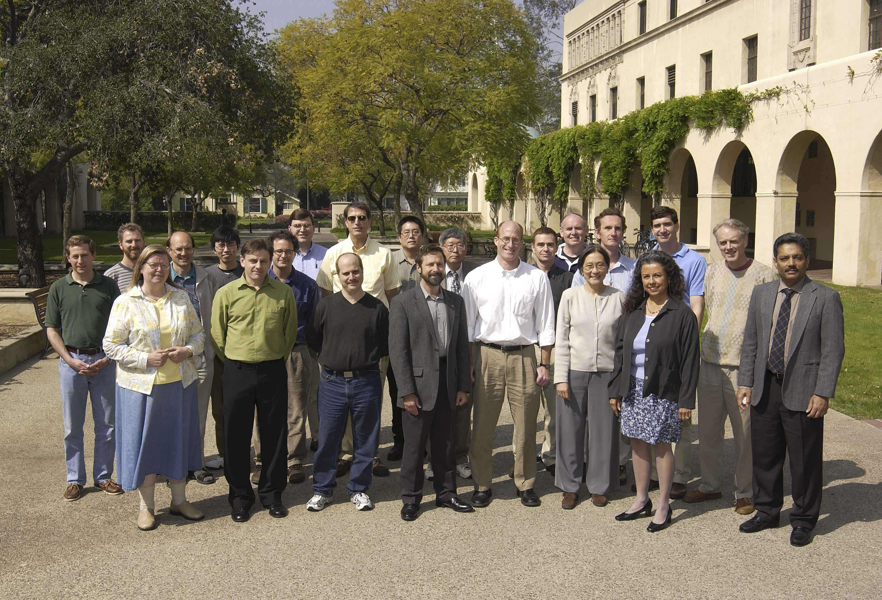 22 people are standing on a wide path at Caltech, in bright sun. A building with arches and vines is behind them. They are all posed in 2 rows and looking at the camera.