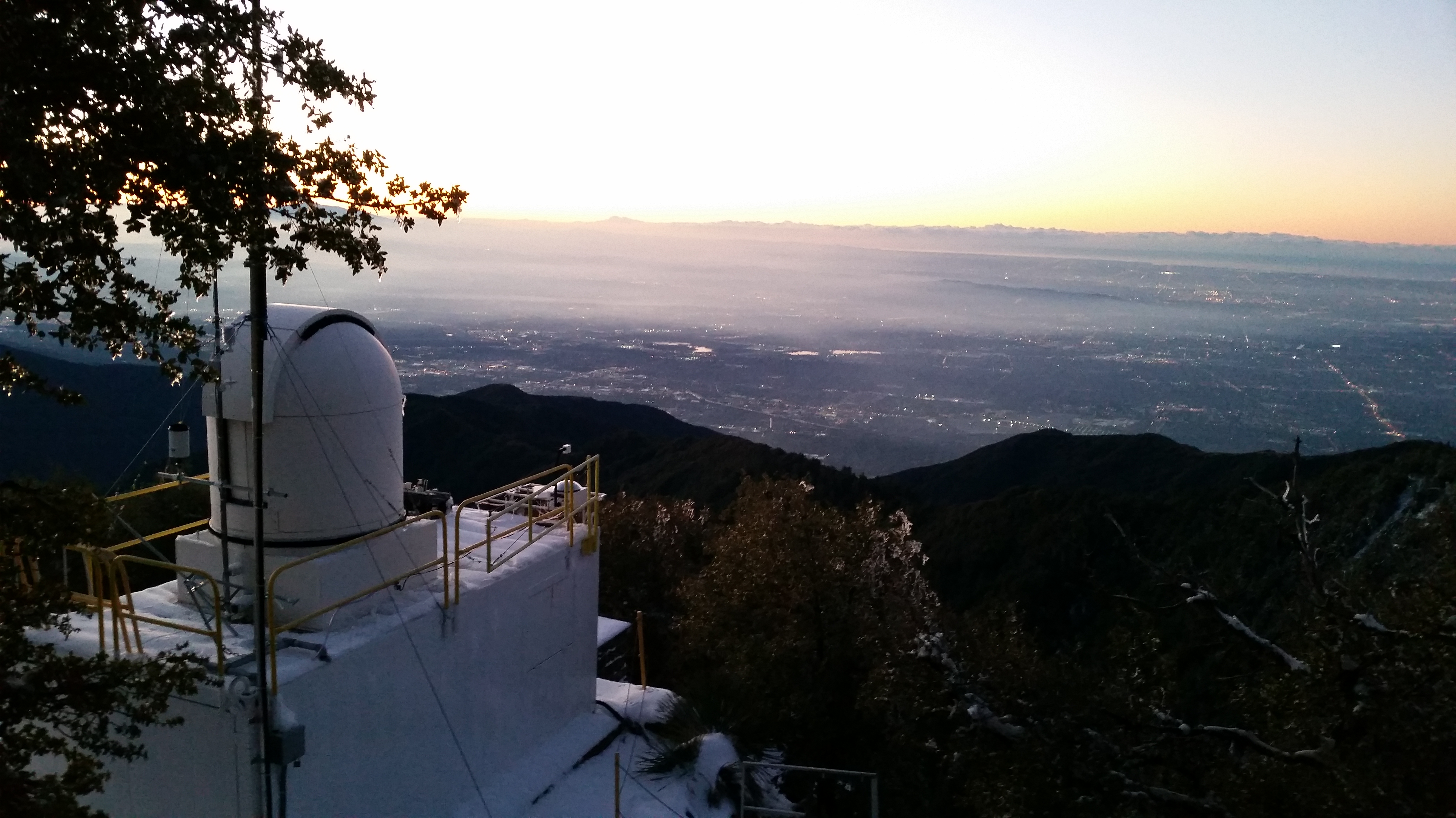 The CLARS facility, a white structure with a tall white dome on top, looks out into Los Angeles valley, the horizon smeared with the blinding white-gold of the sun resting right at the edge. The rest of the valley is shaded in colors of violet and gray, while a misty pall hangs above it. 