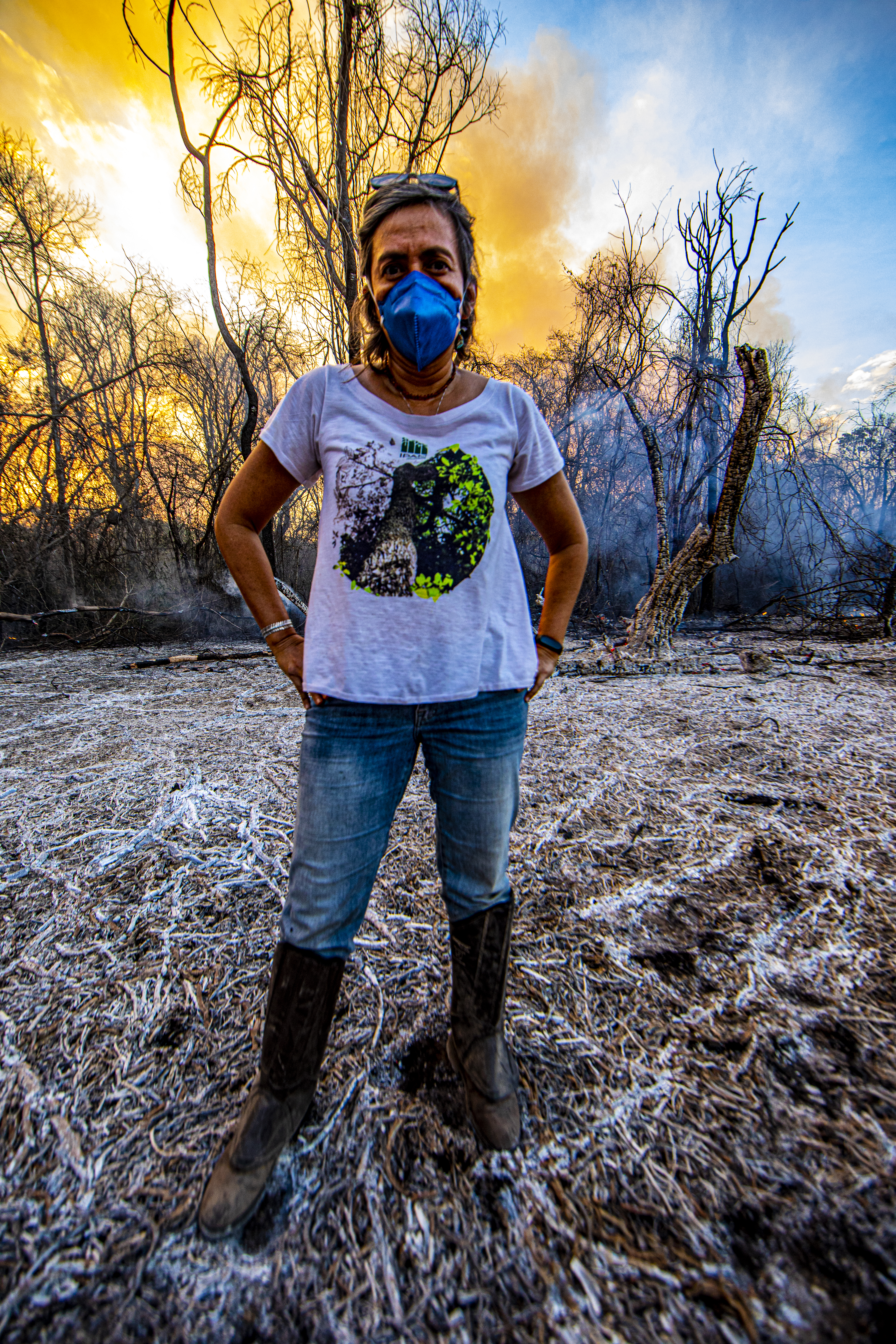 A woman in jeans and a t-shirt, sunglasses perched over her hair, and a mask on stands before charred looking tree skeletons that are silhouetted against a giant blare of orange-white fire right behind her, the flames going up past the edge of the picture