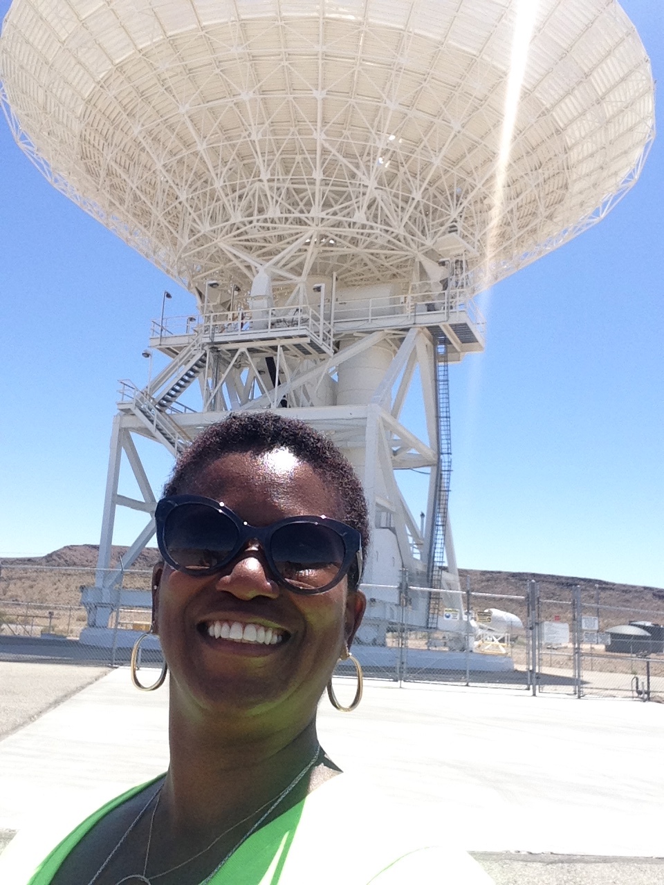 Cozette Parker is standing outside in the desert with a big smile on her face. Tawny-colored hills of sand and dirt stretch out behind her. And a humumgous white antenna with a huge dome and intricate spokes holding it up arises out of the desert right behind her, a streak of brilliant sunlight reflecting against the photo.