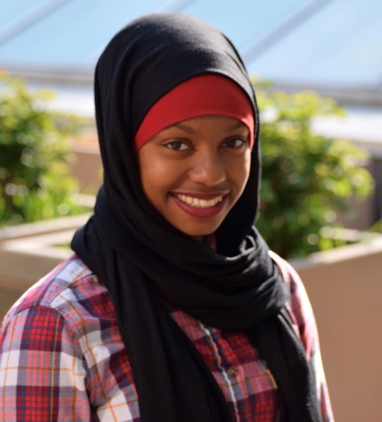 Zainab Ali smiles for a headshot. She is dressed in a red plaid shirt and a red/black hijab combination