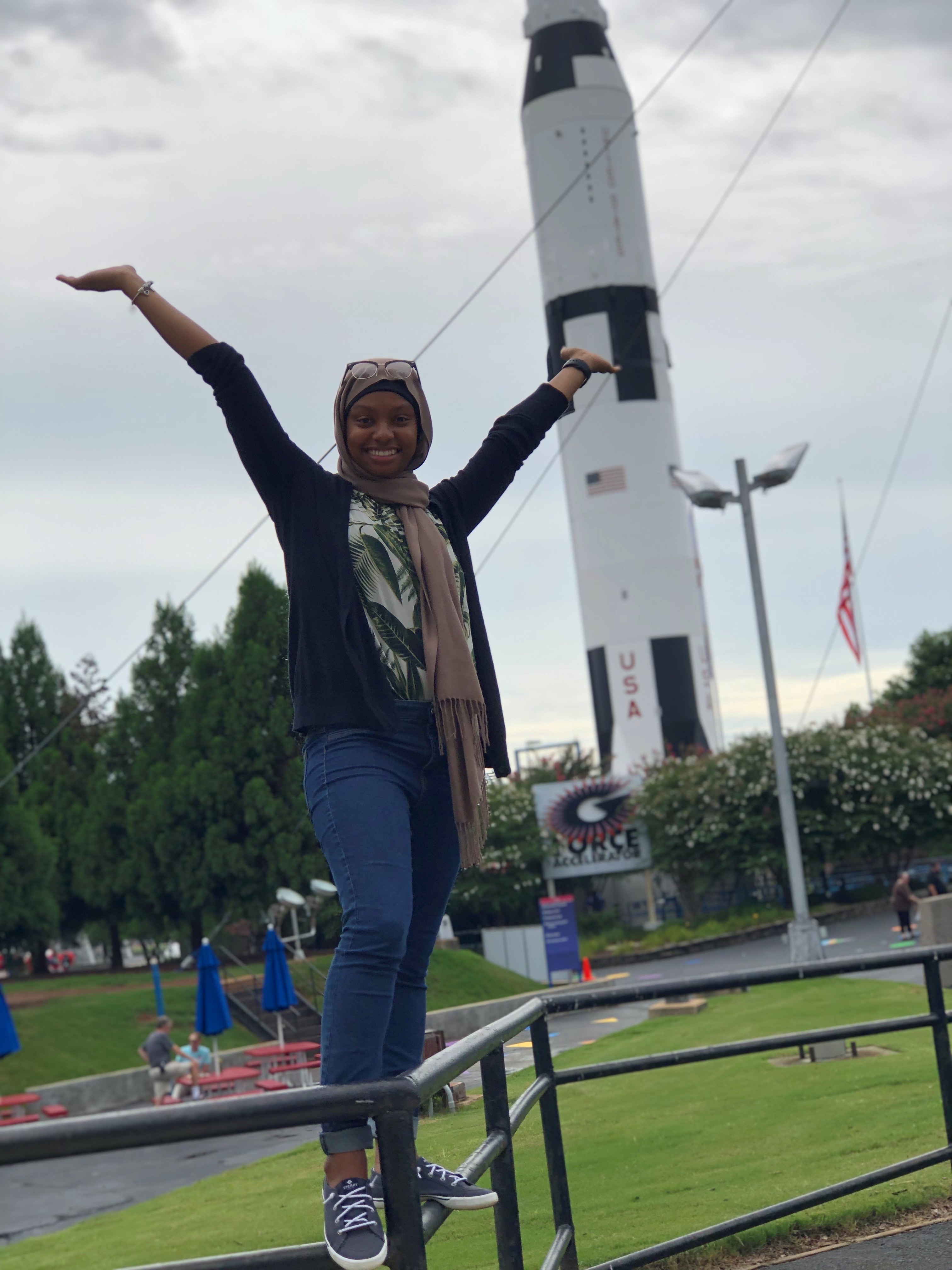 Zainab is posing, with a big smile and arms flung upwards, outdoors before a giant white rocket with the American flag on it. 