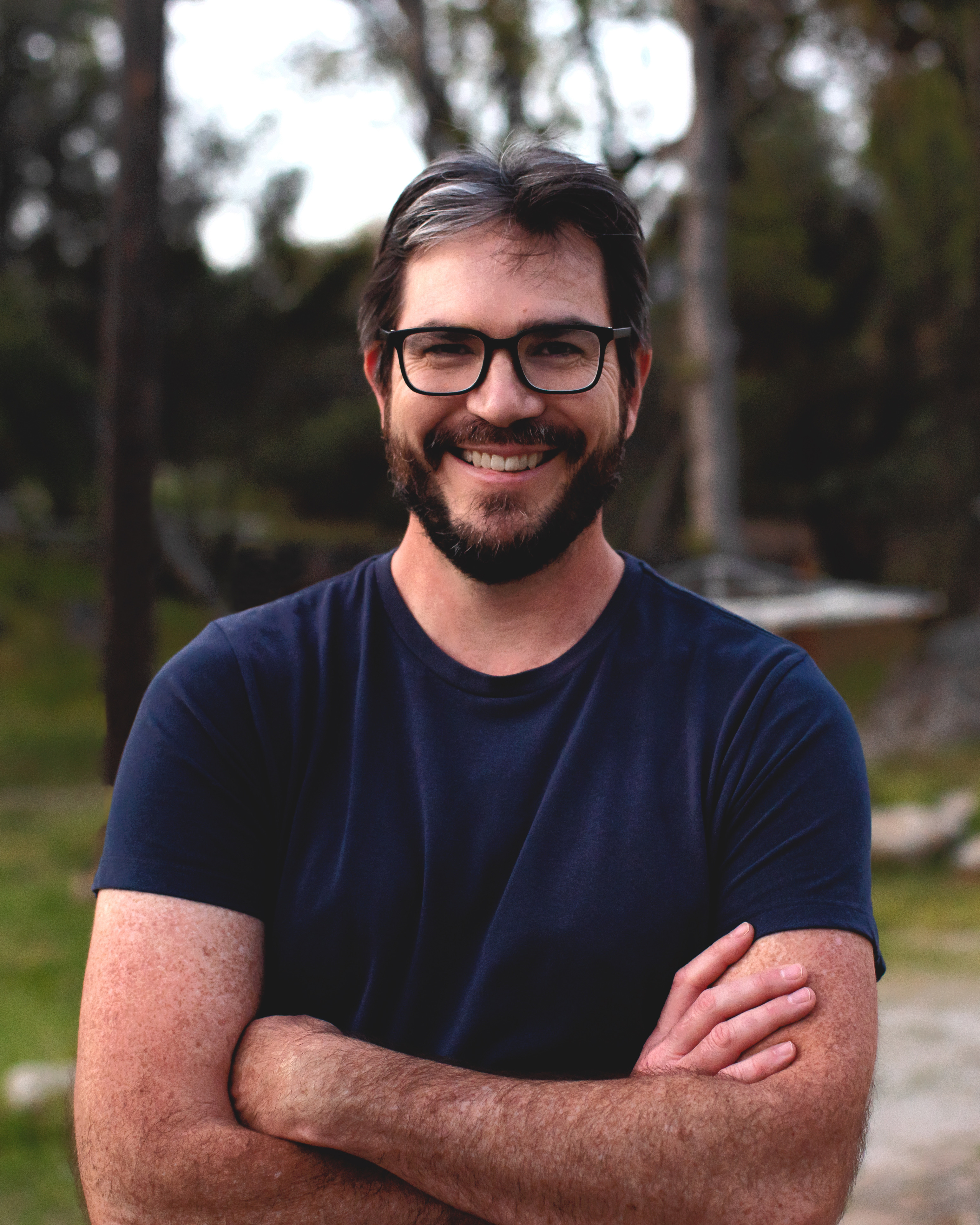 Peter Kalmus, with brown hair, black glasses, brown beard, blue t-shirt, and crossed arms, stands smiling at the camera with a forest behind him