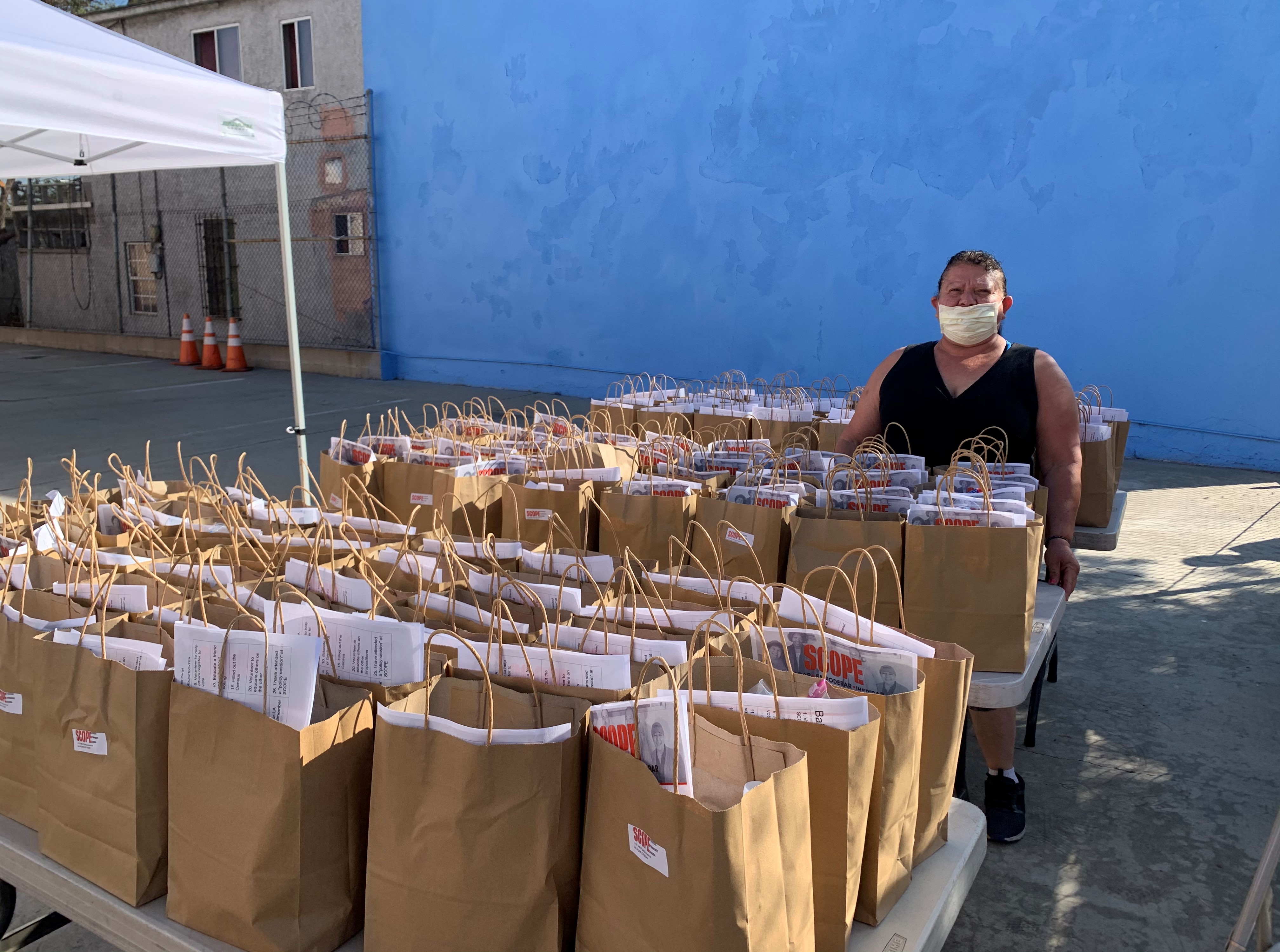 A man in a mask and sleeveless black shirt stands outside, behind a table full of paper bags. Shown sticking out of the paper bags are papers and magazines.