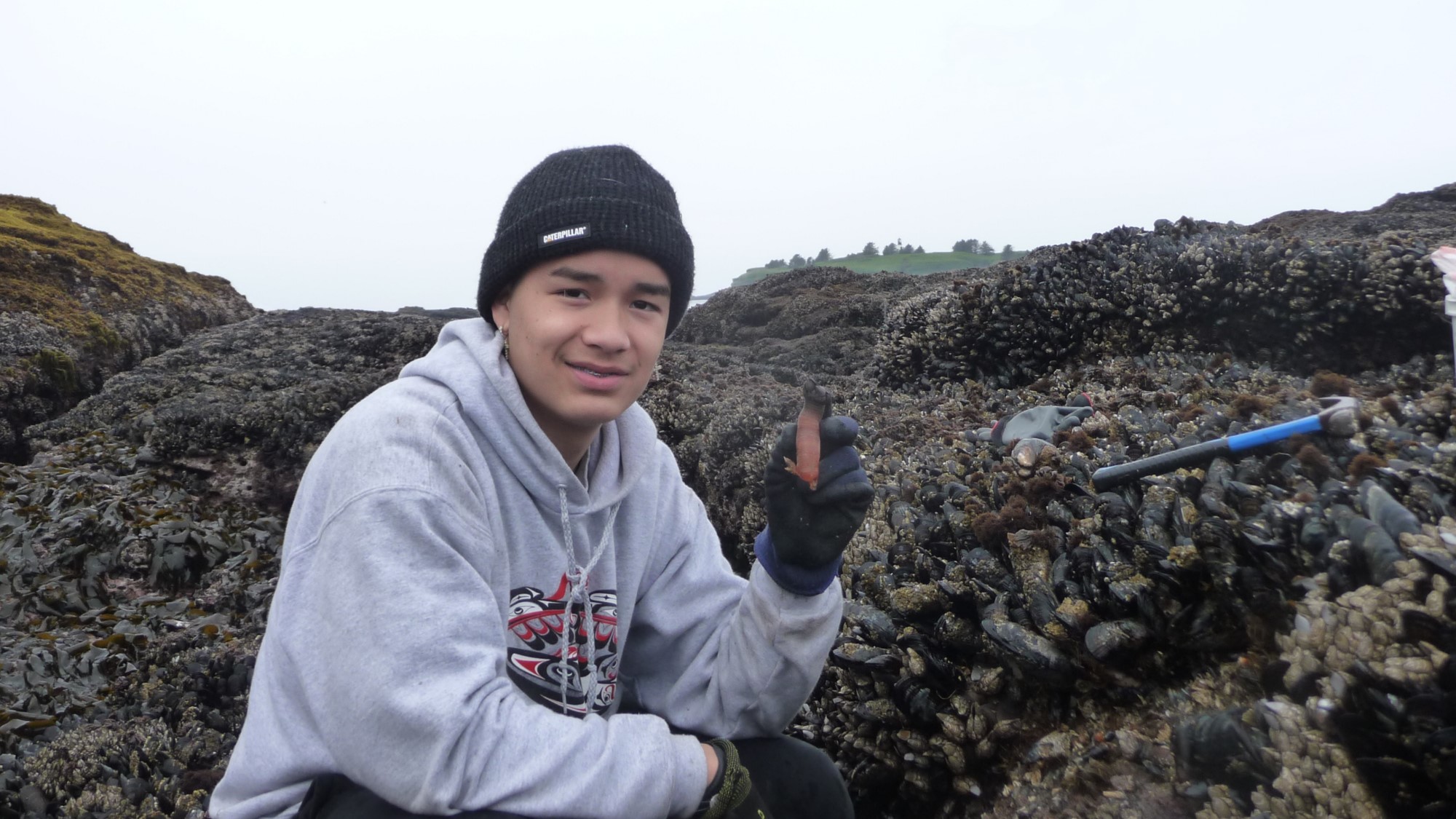 slide 2 - Janine Ledford's son holding up a gooseneck barnacle, while squatting on the rocky coastline, on a cloudy day.