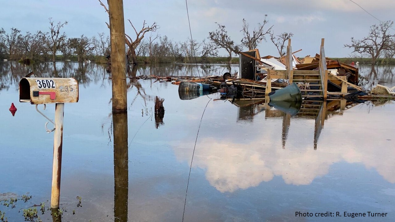 slide 4 - Hurricane Ida's destruction in Louisiana is shown. A lonely mailbox emerges out of flood waters in the foreground, while trees and an electricity pole emerge further back. In the center of the image are the completely disheveled and helter-skelter remains of the home that belonged to the mail box. The only part of it left standing are the stairs going up the front porch.  