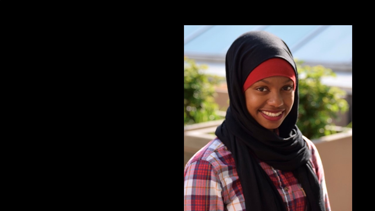slide 3 - Zainab Ali smiles for a headshot. She is dressed in a red plaid shirt and a red/black hijab combination