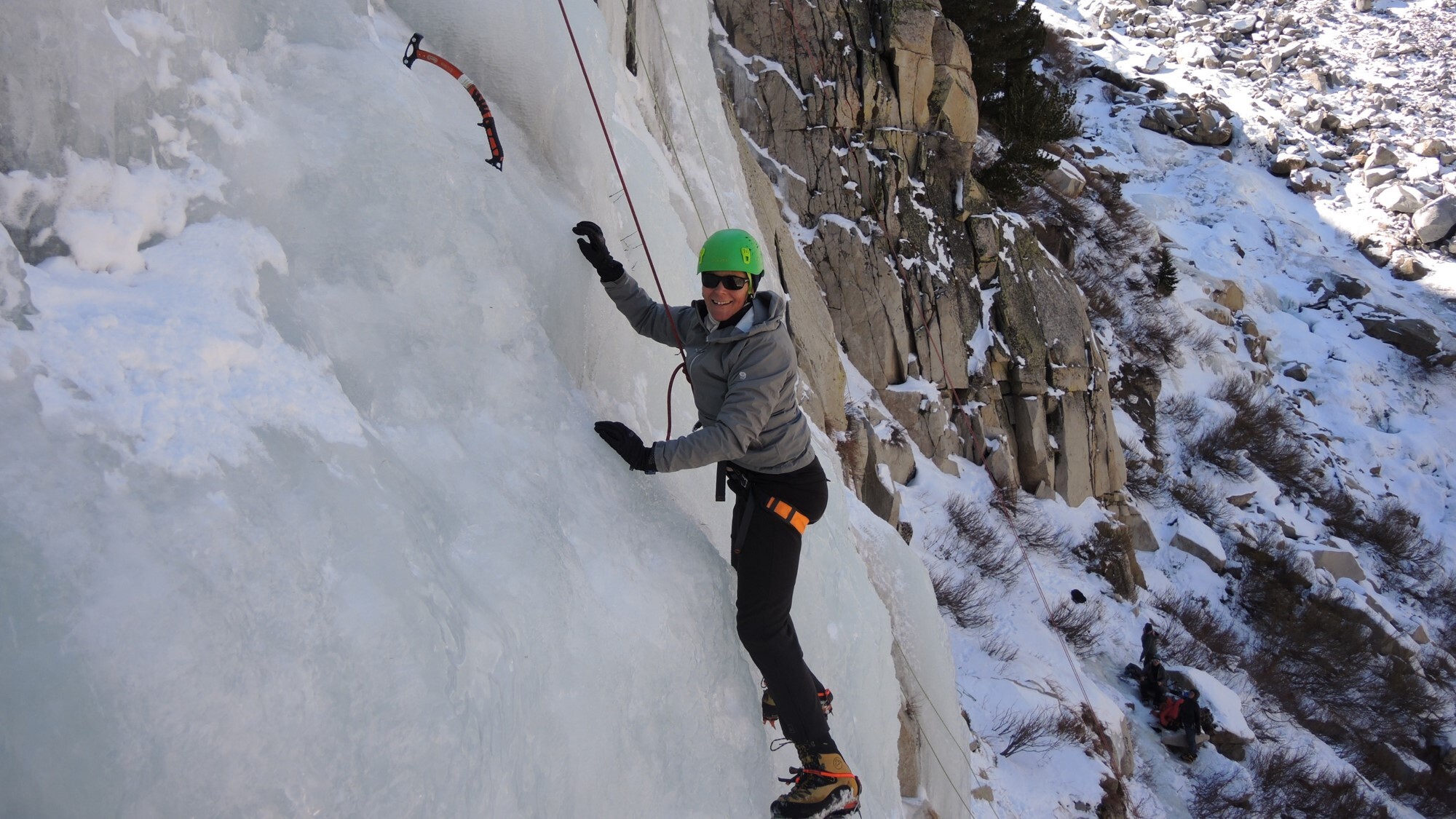 Annmarie Eldering is suspended by rope on a rocky cliffside that is completely covered in a thick layer of ice. She smiles brightly at the camera, dressed in warm gear. 
