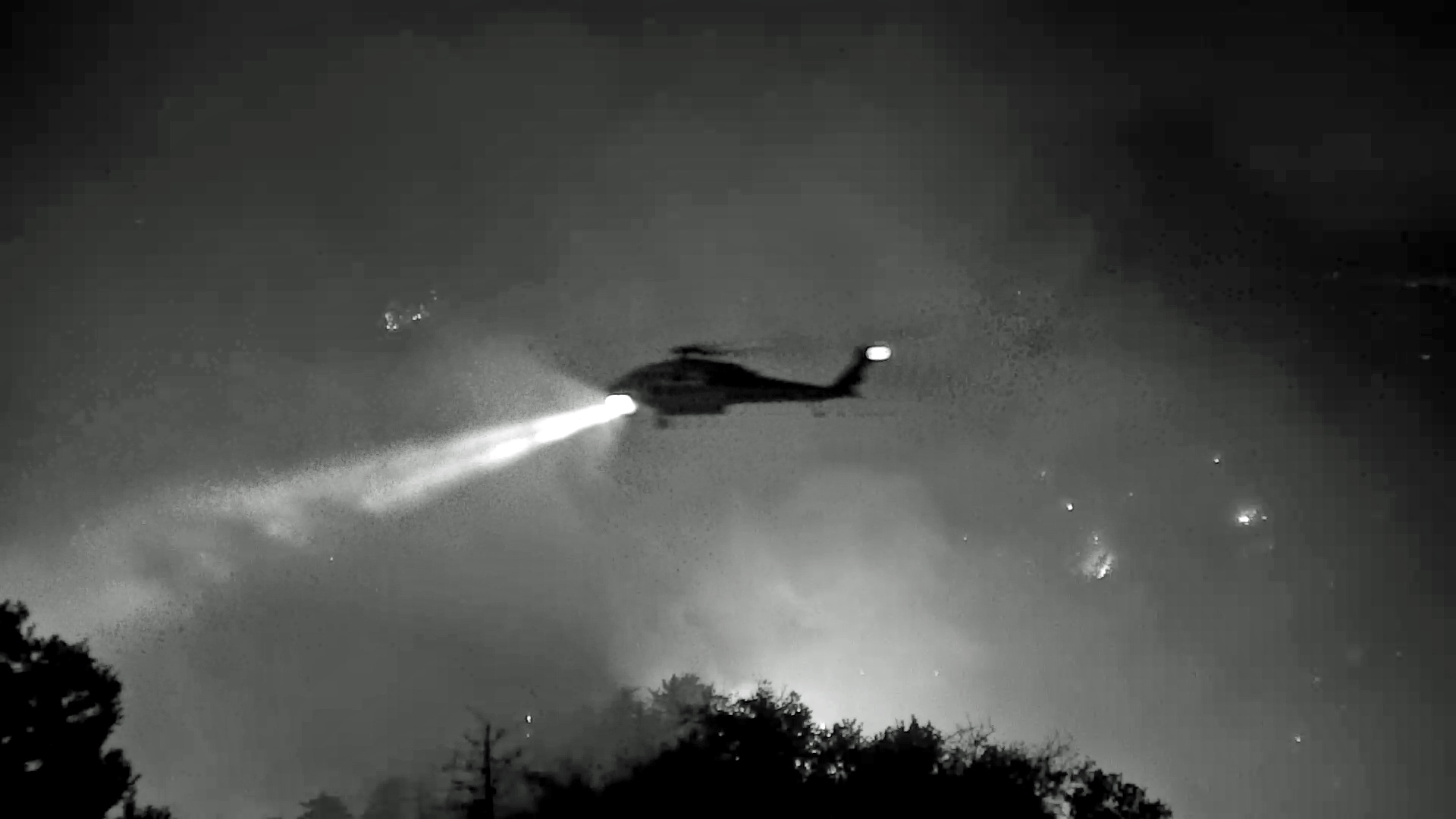 A black and white image taken from a web-camera that shows the dark silhouette of a helicopter against a grainy sky in shades of gray and candlelight. A beacon of bright light erupts from the helicopter and shines down on the tree canopies seen at the very bottom edge of the photo. 